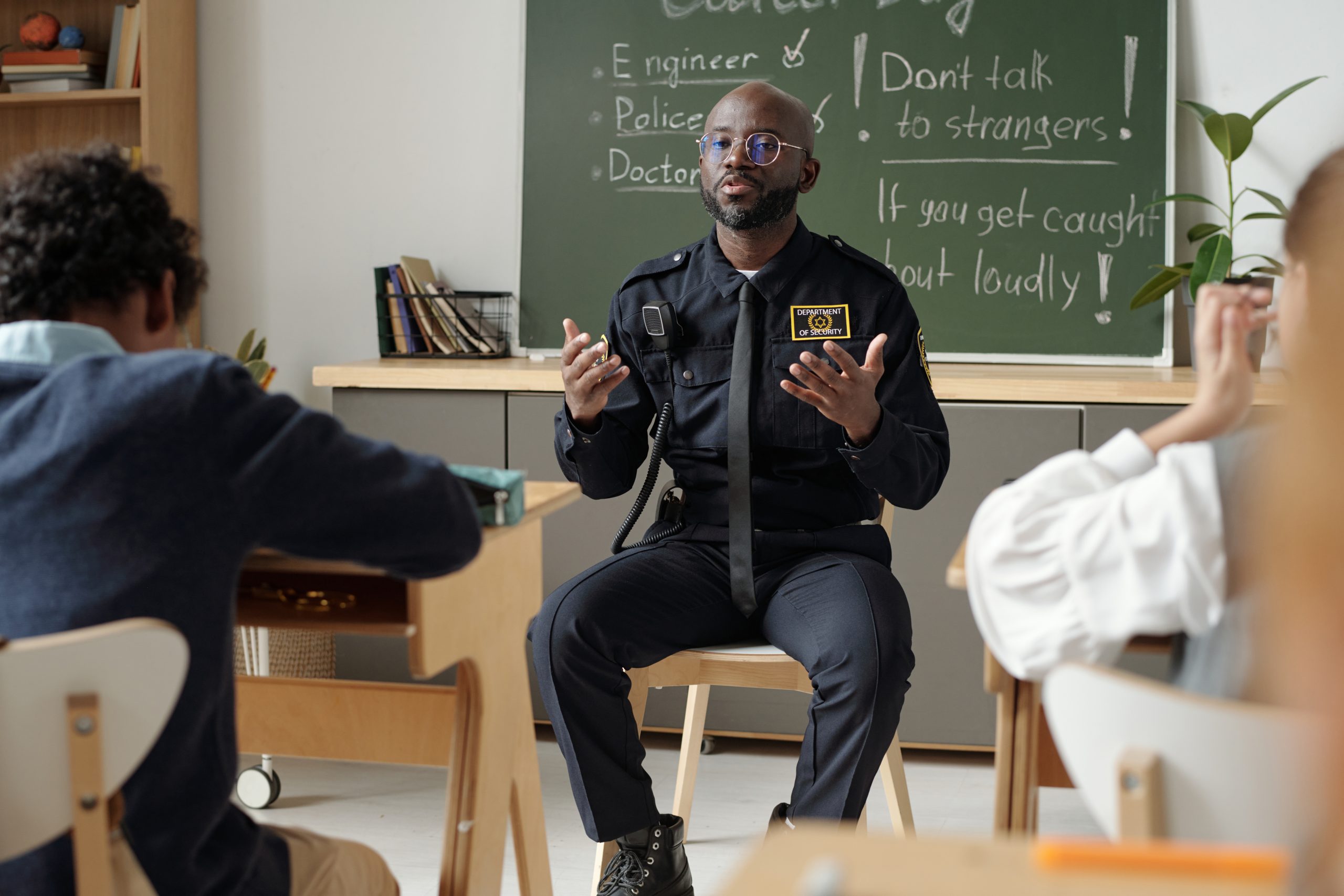 Young confident security guard in uniform sitting in front of diligent children at lesson and speaking about personal safety